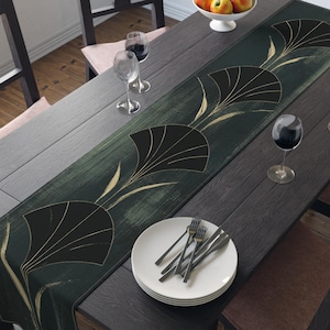 May include: A long rectangular table runner featuring a dark forest green background with a repeating pattern of stylized black and gold fan-shaped botanical motifs. The runner is centered on a dark wooden dining table set with white plates and glasses.