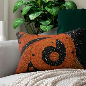 May include: Rectangular decorative pillow with an abstract design in shades of orange, black, and white. The pillow features a spiral pattern and is placed on a gray sofa with a cream-colored blanket.