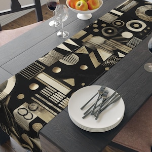 Puede incluir: Un camino de mesa con estampado geométrico en negro y beige se muestra sobre una mesa de madera oscura. El camino de mesa presenta varias formas y patrones. Un cuenco blanco con manzanas, copas de vino y un plato con cubiertos también están sobre la mesa.