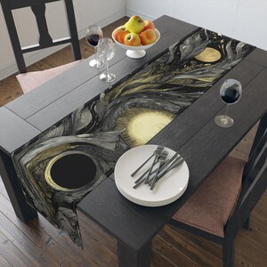 May include: A black dining table set with a decorative table runner featuring a black, gold, and gray celestial design. The table is set with plates, forks, wine glasses, and a bowl of apples. The table is surrounded by black chairs.