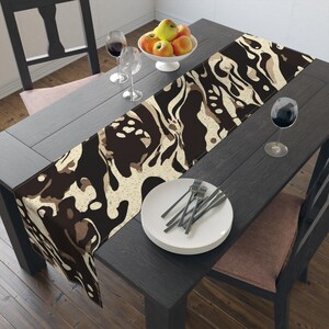 Op de afbeelding: Een decoratieve tafelloper met een bruin en beige abstract patroon. De loper ligt op een donkere houten tafel, aangevuld met een witte kom met appels, wijnglazen en een bord met bestek.