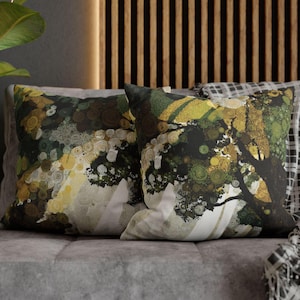 May include: Two decorative throw pillows with a nature-inspired design. The pillows feature a tree and abstract circular patterns in shades of green, yellow, and brown. The pillows are placed on a gray sofa.