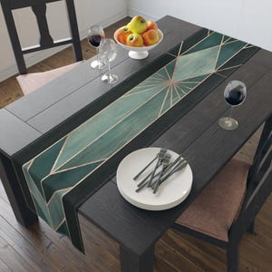 May include: A long rectangular table runner featuring a geometric pattern of dark green and teal triangles accented with thin copper-colored lines. The runner is laid across a dark wooden table set with white plates, silverware, and wine glasses.