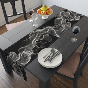 Op de afbeelding: Zwarte tafelloper met een wit, abstract, vloeiend ontwerp. De loper ligt op een donkere houten tafel, gedekt met borden, bestek, wijnglazen en een kom met appels. Zwarte stoelen omringen de tafel.
