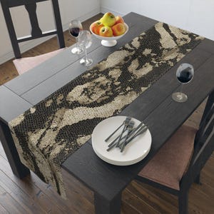 May include: A dark wooden dining table is set with a snakeskin-patterned table runner. White plates, forks, and wine glasses are arranged on the table. A bowl of apples and a glass of red wine complete the setting.