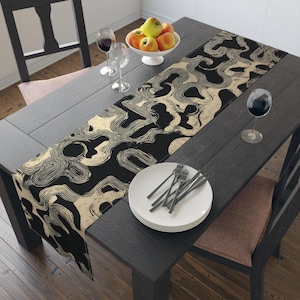 May include: A long rectangular table runner featuring a repeating abstract pattern of flowing, wavy lines in cream against a solid black background. The runner is laid out on a dark wooden table, accompanied by white plates, silverware, and wine glasses.