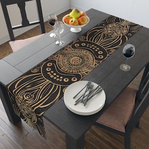 May include: A black table runner with a gold mandala design is displayed on a dark wooden table. The table is set with plates, silverware, wine glasses, and a bowl of apples. The runner features a repeating pattern.