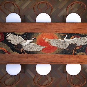 May include: A rectangular wooden dining table with a decorative runner featuring two white cranes with outstretched wings against a black background with a red sun. Six chairs with white seats and wooden frames surround the table.
