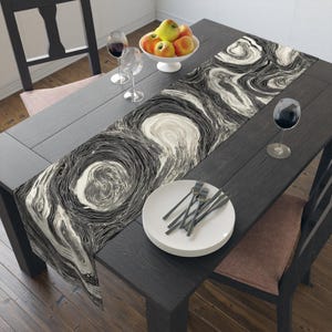 May include: A black and white abstract swirl pattern table runner. The runner is on a dark wood table set with plates, forks, wine glasses, and a bowl of apples. The runner's design resembles marble.