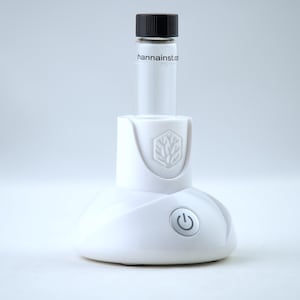 May include: A white electronic device with a small glass vial and a black cap. The vial has the text "hannainst.com". The device has a power button and a logo. The device is likely used for scientific or laboratory purposes.