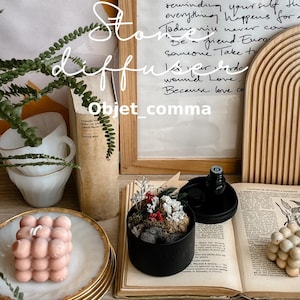 May include: A decorative arrangement with a pink bubble candle, a small potted plant with red and white details, and an open book. The image includes a framed handwritten quote and the text "Objet_comma".
