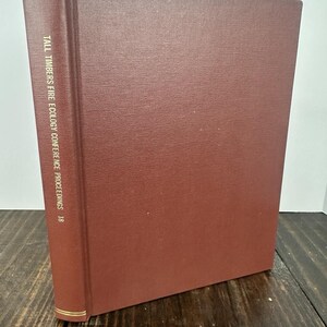 May include: A hardcover book with a textured, reddish-brown cover. The spine features gold accents and the title "TALL TIMBERS FIRE ECOLOGY CONFERENCE PROCEEDINGS 18" in white text. The book is standing upright on a wooden surface.