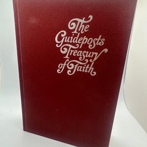 May include: A maroon-colored book titled "The Guideposts Treasury of Faith" with silver lettering. The book has a textured cover and is standing upright. The book's title is written in an elegant, cursive font.