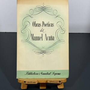 May include: A vintage book titled Obras Poéticas de Manuel Acuña with a cream-colored cover, a light green decorative border, and the text Biblioteca Mundial Sopena at the bottom, resting on a small wooden display stand with the brand name Bookease.