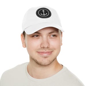May include: White baseball cap with a black and white anchor design. The anchor is inside a circular patch with a rope border. The cap is made of a soft material and has a curved brim.