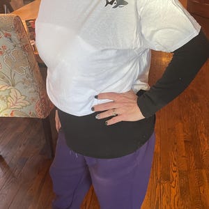 Puede incluir: Camiseta blanca con un peque&ntilde;o gr&aacute;fico de tibur&oacute;n en el pecho, usada sobre una camiseta negra de manga larga. La persona lleva un pantal&oacute;n de ch&aacute;ndal morado y mocasines rosas. El atuendo es informal y c&oacute;modo.