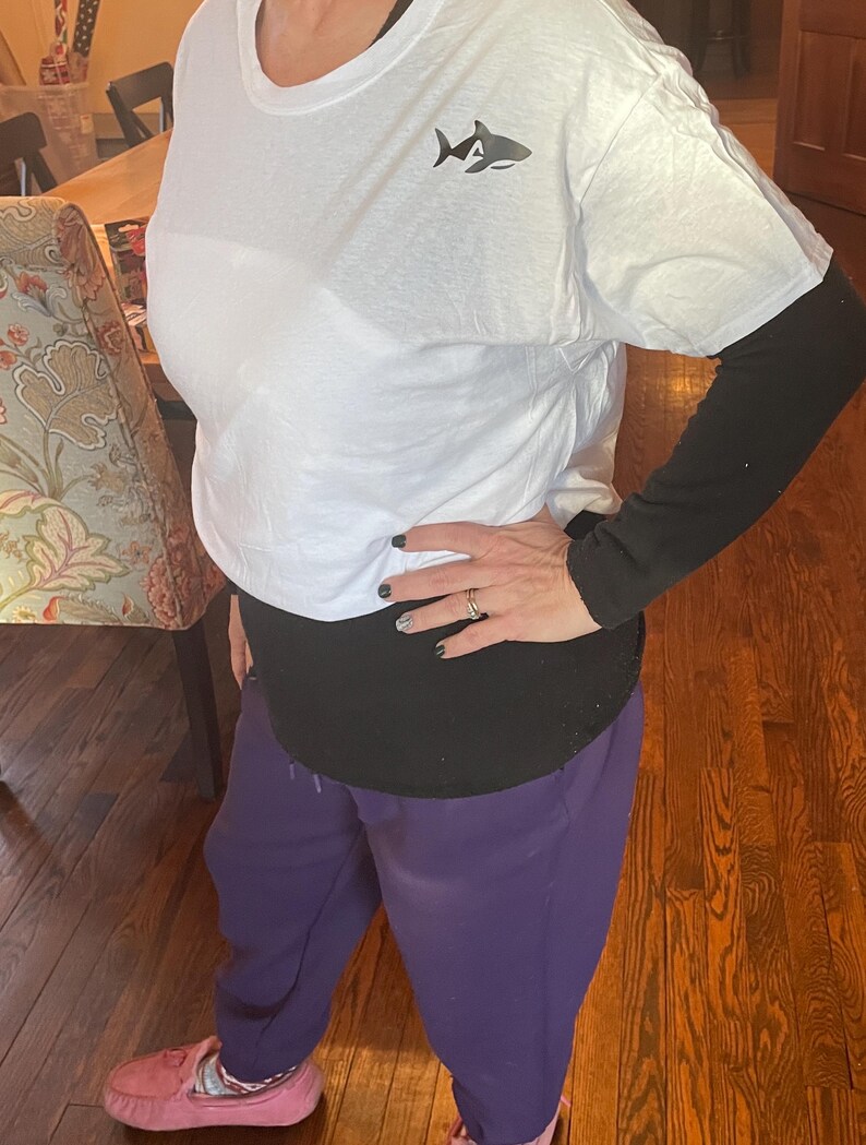 Puede incluir: Camiseta blanca con un peque&ntilde;o gr&aacute;fico de tibur&oacute;n en el pecho, usada sobre una camiseta negra de manga larga. La persona lleva un pantal&oacute;n de ch&aacute;ndal morado y mocasines rosas. El atuendo es informal y c&oacute;modo.