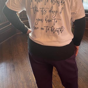 Puede incluir: Camiseta blanca con el texto "If sharks have to move to breathe, then it's okay if you have to move on to breathe" en escritura negra. La persona lleva mangas negras y pantalones morados.