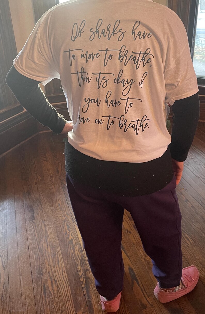 Puede incluir: Camiseta blanca con el texto "If sharks have to move to breathe, then it's okay if you have to move on to breathe" en escritura negra. La persona lleva mangas negras y pantalones morados.