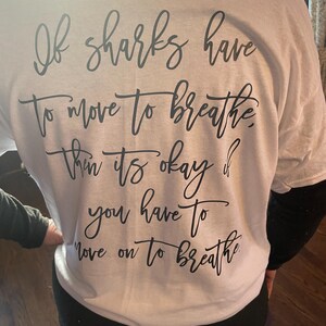 Puede incluir: Camiseta blanca con texto negro en cursiva. El texto dice: "Si los tiburones tienen que moverse para respirar, entonces est&aacute; bien si tienes que seguir adelante para respirar."