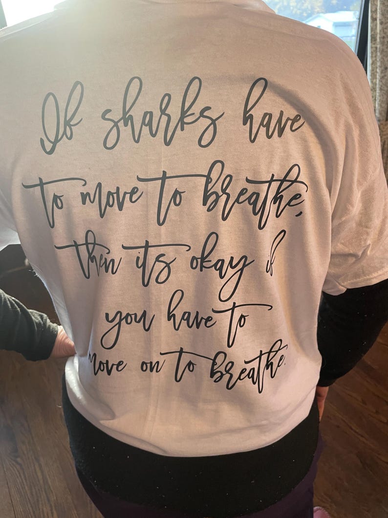 Puede incluir: Camiseta blanca con texto negro en cursiva. El texto dice: "Si los tiburones tienen que moverse para respirar, entonces est&aacute; bien si tienes que seguir adelante para respirar."