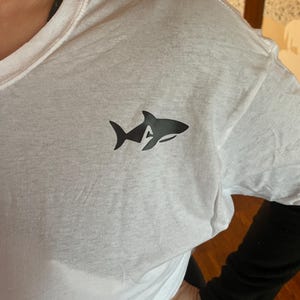Puede incluir: Camiseta blanca con un gr&aacute;fico de tibur&oacute;n negro en el pecho. El dise&ntilde;o del tibur&oacute;n incorpora la letra "A" en su silueta. La camiseta est&aacute; hecha de un material suave y ligero, adecuado para ropa casual.