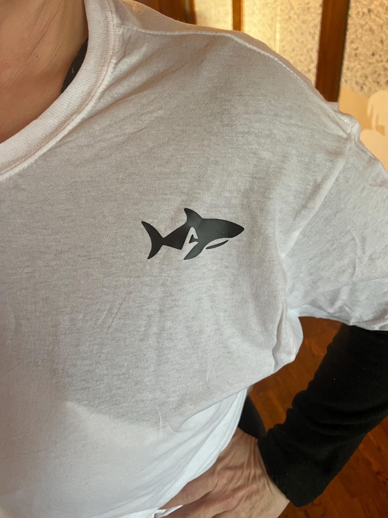 Puede incluir: Camiseta blanca con un gr&aacute;fico de tibur&oacute;n negro en el pecho. El dise&ntilde;o del tibur&oacute;n incorpora la letra "A" en su silueta. La camiseta est&aacute; hecha de un material suave y ligero, adecuado para ropa casual.