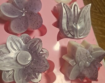 May include: A pink silicone mould filled with handmade soaps in various floral shapes. The soaps are lavender and white, with speckled details. The mould includes flower, tulip, and circular designs.