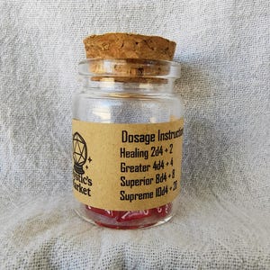 May include: A small glass jar with a cork stopper, filled with red dice. The jar has a label with the text "Dosage Instructions" and other text. The jar is on a white, textured surface.