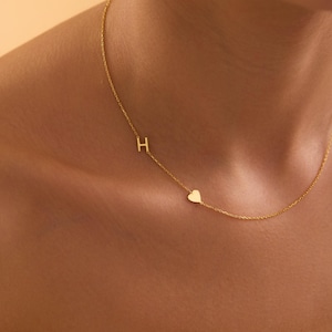 May include: A delicate gold necklace featuring the letter "H" and a small heart pendant. The necklace is worn around the neck, showcasing the minimalist design and elegant simplicity of the jewellery.