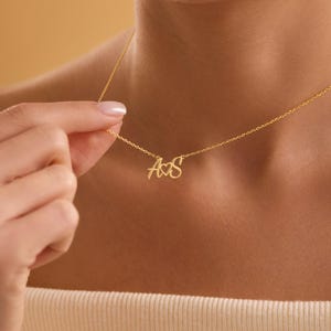 May include: A delicate gold necklace featuring the initials "A&S" in a cursive font. The pendant hangs from a fine chain, creating an elegant and personalized accessory. The necklace is shown against a warm, neutral background.