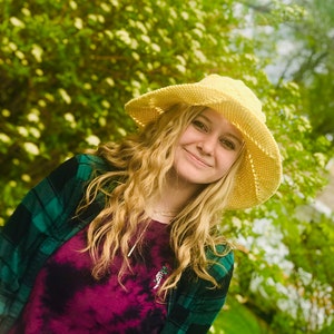 May include: A young woman with long blonde hair is wearing a yellow crochet sun hat and a green and black plaid shirt over a purple tie-dye shirt. She is smiling at the camera.