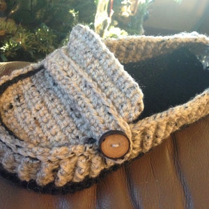 May include: A gray and black crocheted slipper with a brown button. The slipper is made with a textured yarn and has a black trim around the edge.