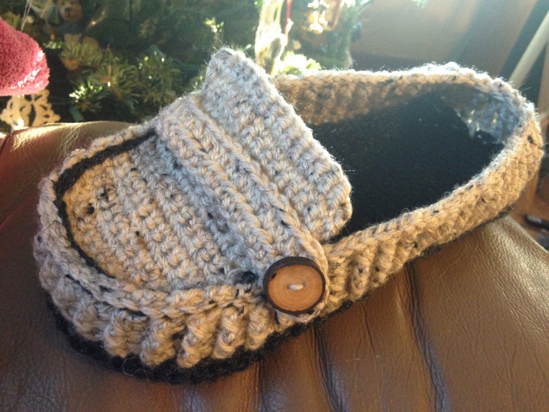 May include: A gray and black crocheted slipper with a brown button. The slipper is made with a textured yarn and has a black trim around the edge.