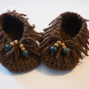 May include: A pair of dark brown crocheted booties featuring a fringe detail around the opening and a decorative tie at the front. Each bootie is accented with two light wooden beads and one dark teal bead threaded onto the yarn ties at the center.