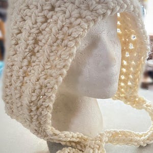 May include: Cream-colored, hand-crocheted hood with a textured stitch and silver metallic thread accents. The hood features a long, braided tie and is displayed on a mannequin head. This cozy accessory is perfect for cold weather.