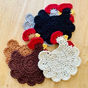 May include: Four crocheted rooster-shaped coasters in different colors: brown, white, black, and gray. The coasters have red and yellow accents.