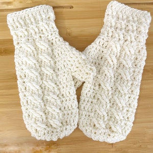 May include: A pair of cream-colored, hand-crocheted mittens. The mittens feature a textured cable knit design and are made of a soft, warm yarn. The mittens are shown on a wooden surface.