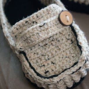 May include: A close-up of a beige crocheted slipper with a black trim and a wooden button. The slipper is made with a chunky yarn and has a textured surface.