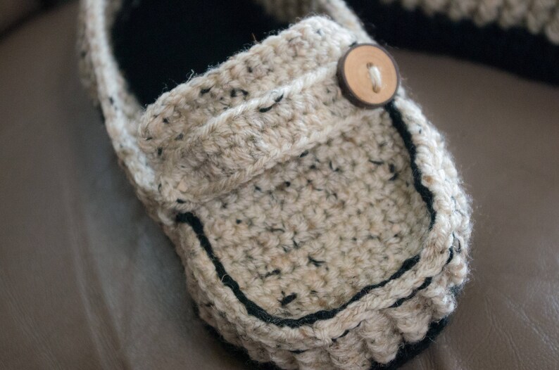 May include: A close-up of a beige crocheted slipper with a black trim and a wooden button. The slipper is made with a chunky yarn and has a textured surface.