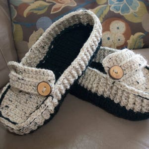 May include: A pair of crocheted slippers with a beige and black color scheme. Each slipper has a wooden button on the top.