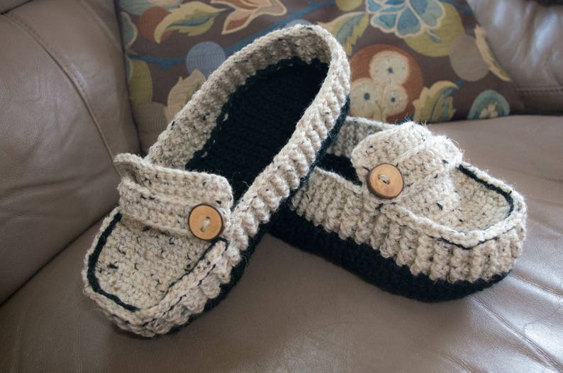 May include: A pair of crocheted slippers with a beige and black color scheme. Each slipper has a wooden button on the top.