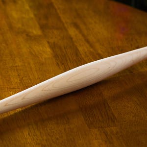 May include: A wooden rolling pin, approximately 18 inches long, with a smooth, light brown finish. The rolling pin is lying on a wooden surface.