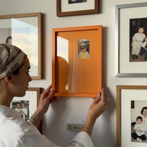 Puede incluir: Un marco de fotos naranja con una foto pequeña en su interior, sostenido contra una pared blanca. Otras fotos enmarcadas de varios tamaños y estilos también están en la pared. Un nivel está montado debajo del marco.