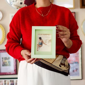May include: A rectangular light green picture frame holding a photograph of a person in a swimsuit sitting on a sandy beach. The frame has a smooth, matte finish and a clean, simple border design, showcasing a vintage-style image inside.