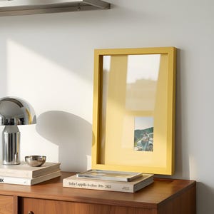 May include: A yellow rectangular picture frame with a small photograph inside. A silver lamp, a small silver bowl, and books are on a wooden surface. The frame is leaning against a white wall.