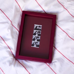 May include: A rectangular deep burgundy picture frame featuring a centred vertical strip of four black and white photo booth images. The photos depict two individuals and a small dog in various poses, displayed against a matching burgundy mount board.