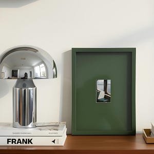May include: A polished chrome mushroom-shaped table lamp reflects the room, placed on books. A framed picture with a green border displays a small image of a train interior. The items are on a wooden surface against a white wall.