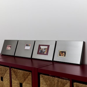 Peut inclure: Quatre cadres photo carrés en métal argenté brossé disposés en rangée. Chaque cadre présente une large bordure plate et une petite fenêtre rectangulaire centrée affichant une photographie unique. Les cadres ont des passe-partouts de couleurs variées.