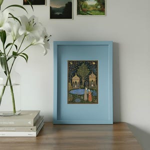 May include: A framed artwork in a light blue frame. The artwork shows a detailed scene with two figures near a pond, surrounded by trees and ornate structures. The artwork is displayed on a wooden surface, with a vase of white lilies and books nearby.